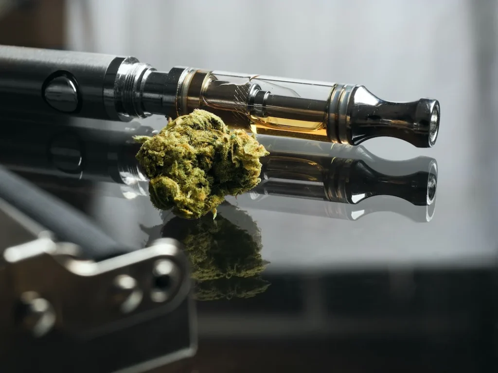 Two vape cartridges and a marijuana bud on a dark reflective surface